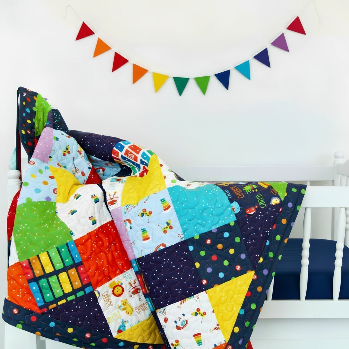 Fisher Price patchwork quilt draped over white crib navy sheets rainbow bunting nursery Riley Blake novelty kids fabric baby quilt